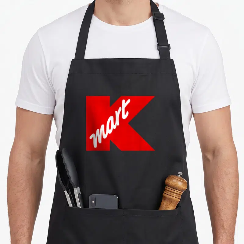 Department kmart apron - Large red letter K background with white mart script written diagonally across the front — shown on a black cotton canvas apron against a white background