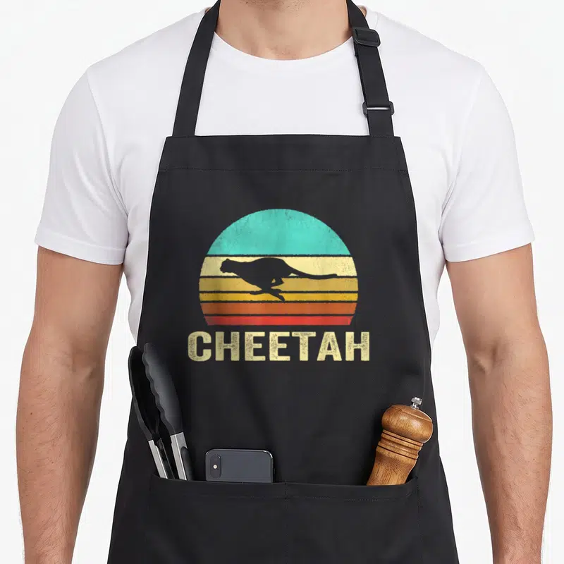 Sunset cheetah apron - Grey cheetah silhouette leaping over retro striped sun with CHEETAH text below — shown on a black cotton canvas apron against a white background