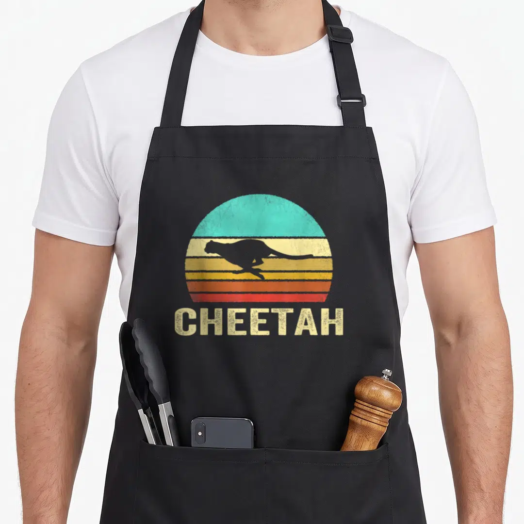 Sunset cheetah apron - Grey cheetah silhouette leaping over retro striped sun with CHEETAH text below — shown on a black cotton canvas apron against a white background