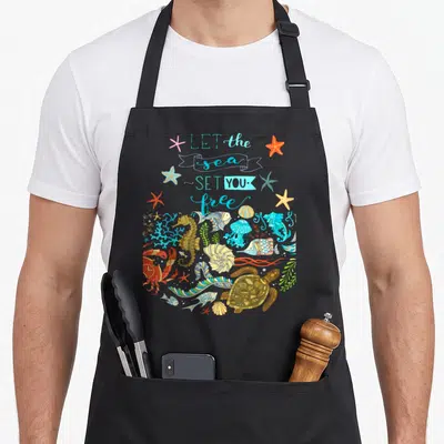 Sea marine apron - Cartoon sea creatures and starfish below LET the sea SET YOU free text in blue and orange — shown on a black cotton canvas apron against a white background