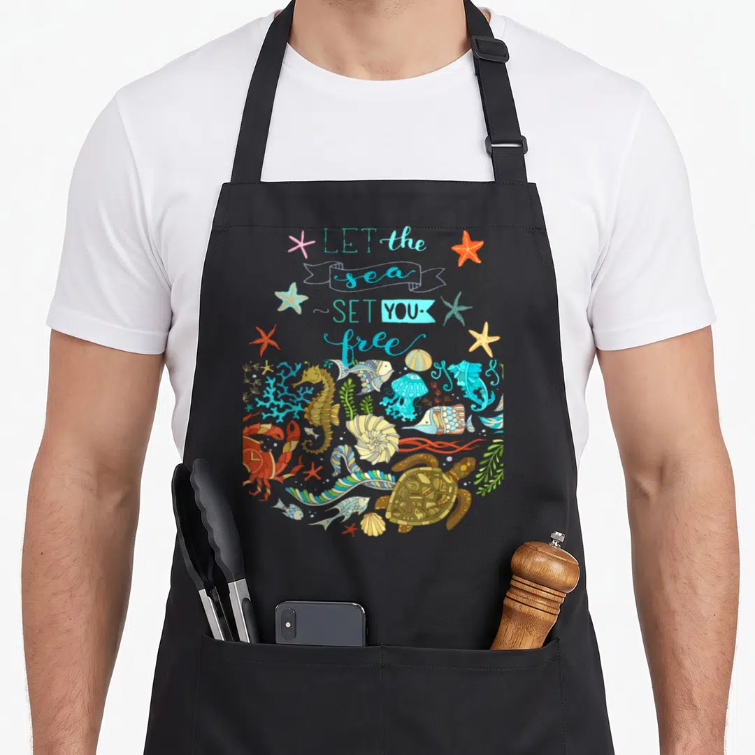 Sea marine apron - Cartoon sea creatures and starfish below LET the sea SET YOU free text in blue and orange — shown on a black cotton canvas apron against a white background