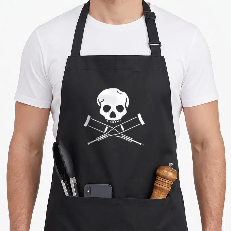 Jackass skull apron - White skull graphic above two crossed crutches in minimalist style — shown on a black cotton canvas apron against a white background