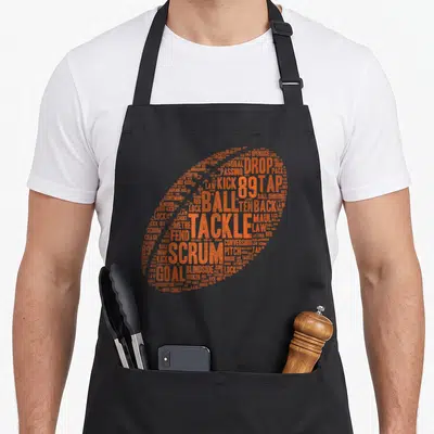 Rugby pride apron - Orange rugby ball formed by text including scrum, tackle, goal, and law — shown on a black cotton canvas apron against a white background