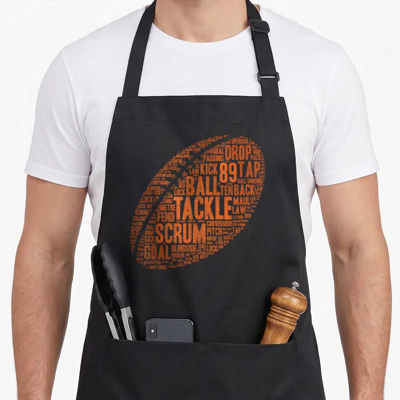 Rugby pride apron - Orange rugby ball formed by text including scrum, tackle, goal, and law — shown on a black cotton canvas apron against a white background
