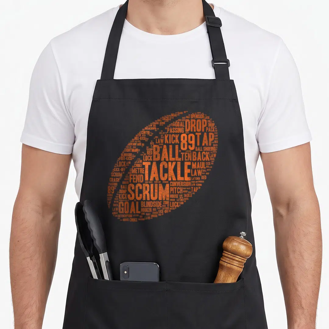 Rugby pride apron - Orange rugby ball formed by text including scrum, tackle, goal, and law — shown on a black cotton canvas apron against a white background
