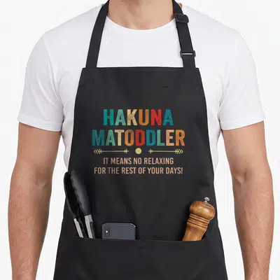 Hakuna tea quotes apron - HAKUNA MATODDLER text in teal orange red with decorative line and circle separating — shown on a black cotton canvas apron against a white background