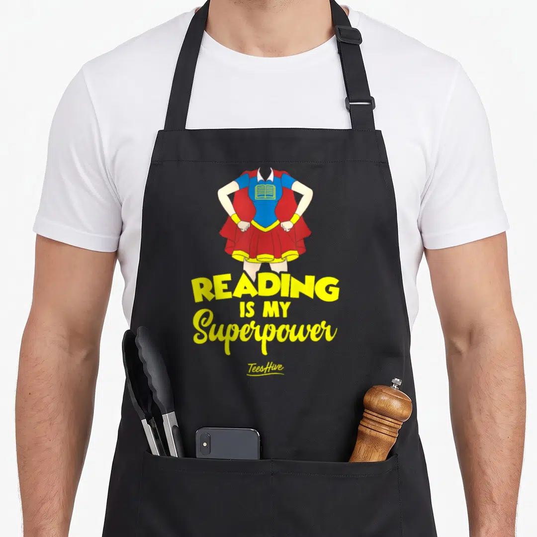 Reading writing lover apron - Headless female superhero torso with red cape and blue top holding a yellow book above — shown on a black cotton canvas apron against a white background