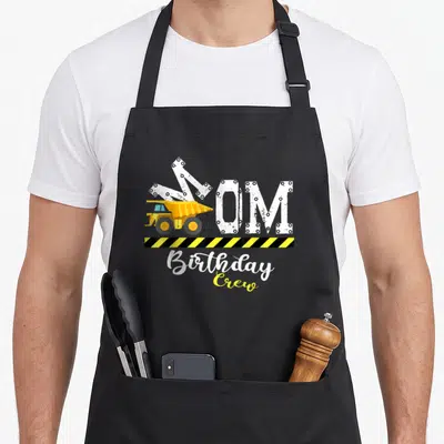 Mother building aprons - Yellow dump truck overlapping white MOM text above yellow and black hazard stripe — shown on a black cotton canvas apron against a white background
