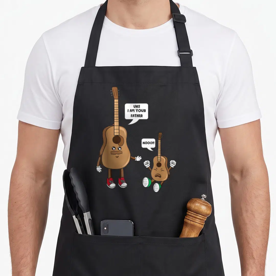 Uke acoustics nerd apron - Cartoon guitar saying UKE I AM YOUR FATHER to a fleeing ukulele — shown on a black cotton canvas apron against a white background