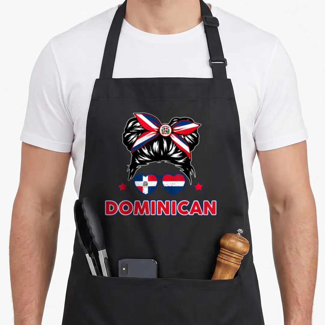 Republica hispanic aprons - Cartoon messy bun with flag bow, heart sunglasses, two stars, and DOMINICAN text — shown on a black cotton canvas apron against a white background