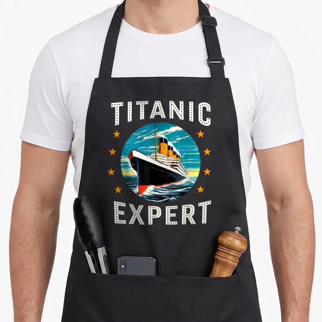 Titanic line apron - Retro ship in blue circle flanked by three orange stars with TITANIC EXPERT text in white dots — shown on a black cotton canvas apron against a white background