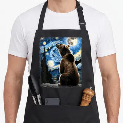Artistic van gogh apron - Brown bear facing swirling blue night sky with moon stars and cypress tree watercolor — shown on a black cotton canvas apron against a white background