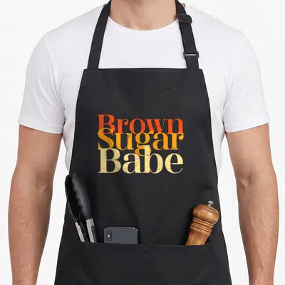 History sugar apron - Stacked Brown Sugar Babe text in red-orange, orange, and beige distressed fonts — shown on a black cotton canvas apron against a white background