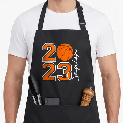 Funny senior class player apron - Orange 2023 basketball graphic with vertical white senior text on black background — shown on a black cotton canvas apron against a white background