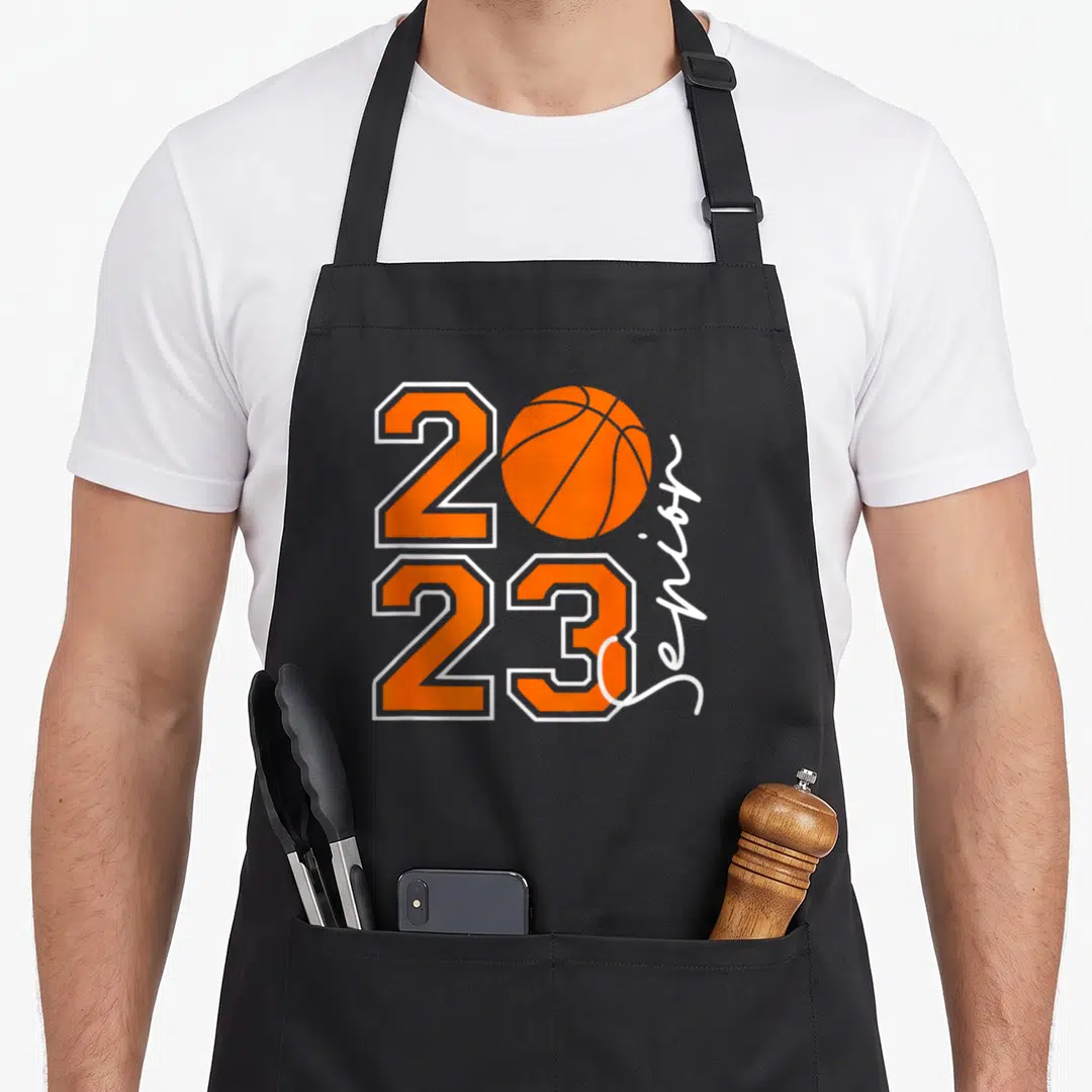 Funny senior class player apron - Orange 2023 basketball graphic with vertical white senior text on black background — shown on a black cotton canvas apron against a white background