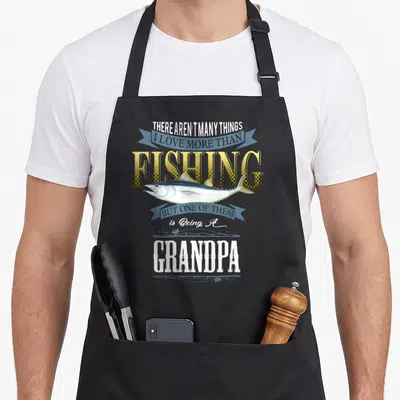 Grandpa fishing aprons - Stacked text about loving fishing and being a grandpa with a fish and ribbons in yellow blue — shown on a black cotton canvas apron against a white background