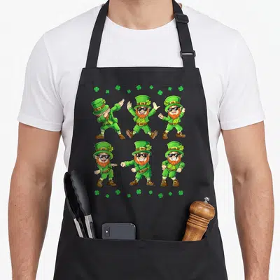 Dancing st patrick day aprons - Six cartoon leprechauns in green and orange performing dab and floss dance poses — shown on a black cotton canvas apron against a white background
