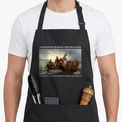 Winning wars classic apron - Washington Crossing the Delaware painting with date text and two front pockets — shown on a black cotton canvas apron against a white background