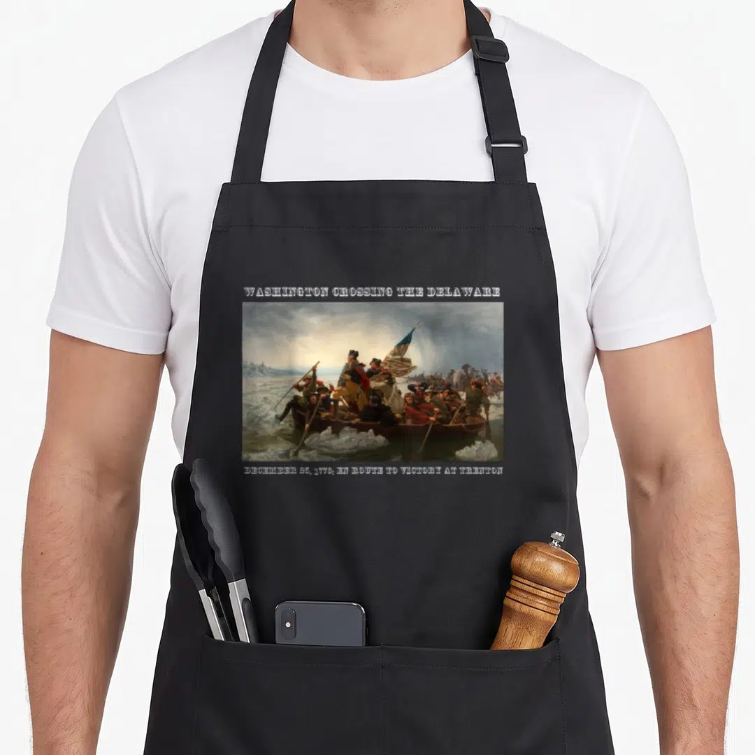 Winning wars classic apron - Washington Crossing the Delaware painting with date text and two front pockets — shown on a black cotton canvas apron against a white background