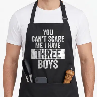 Cute scare dad and son aprons - White stacked text You Can't Scare Me I Have Three Boys distressed style — shown on a black cotton canvas apron against a white background