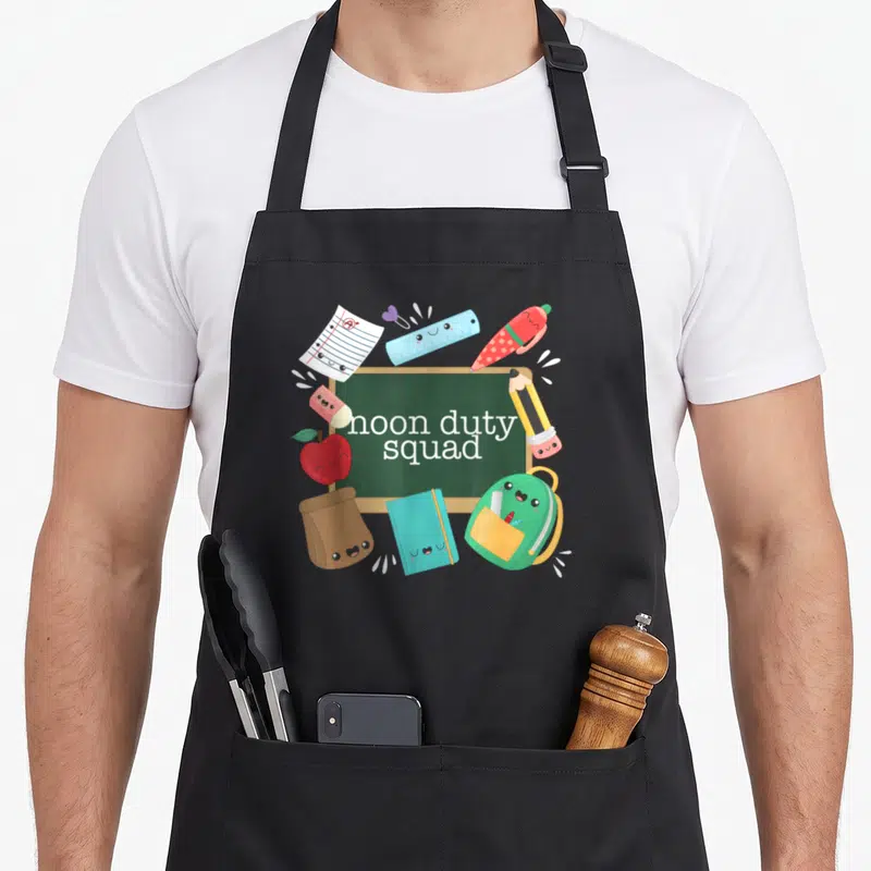 Noon kawaii apron - Chalkboard with smiling ruler, pencil, backpack, apple, notebook, paper, eraser, and bag — shown on a black cotton canvas apron against a white background