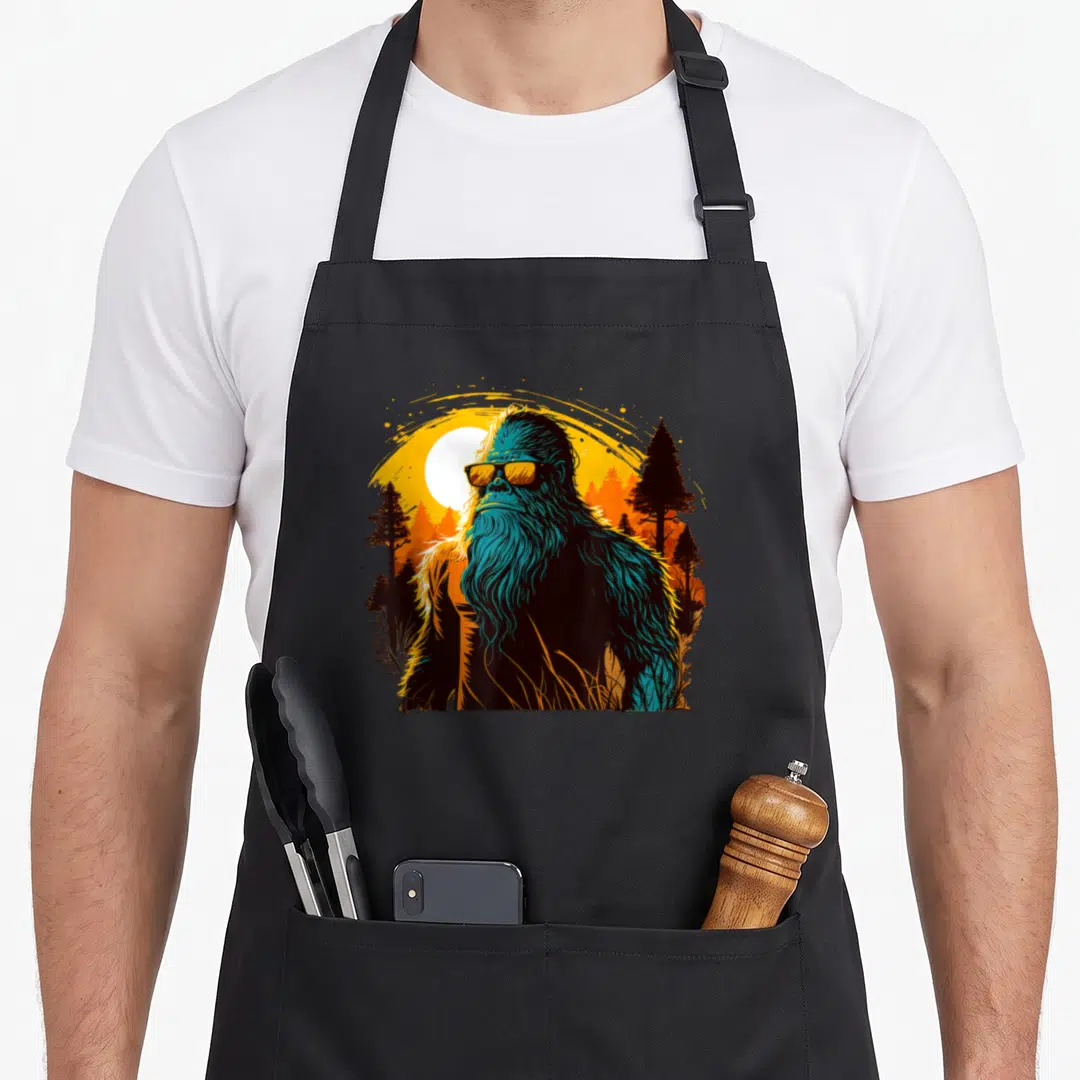 Sunset yeti apron - Retro Bigfoot in teal and orange sunglasses standing in a pine forest under a yellow sun — shown on a black cotton canvas apron against a white background