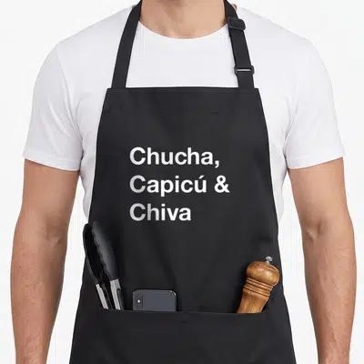 Dominoe spanish aprons - White text reading Chucha Capicú Chiva stacked vertically — shown on a black cotton canvas apron against a white background