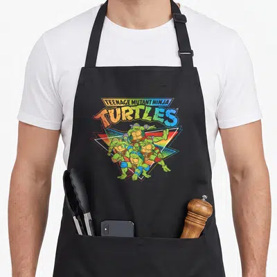 Pride gradient rainbow apron - Stacked TMNT text above four turtles over rainbow triangle in cartoon style — shown on a black cotton canvas apron against a white background