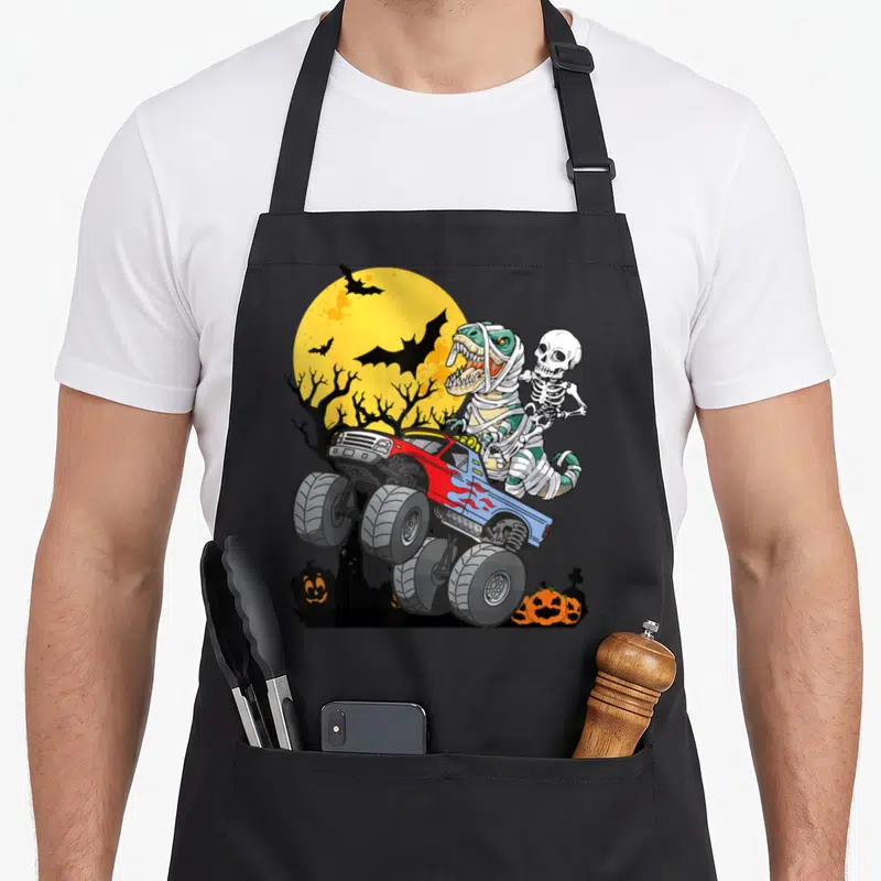 Pumpkin truck apron - Cartoon monster truck with mummy dinosaur skeleton riding under yellow moon with bats and pumpkins — shown on a black cotton canvas apron against a white background