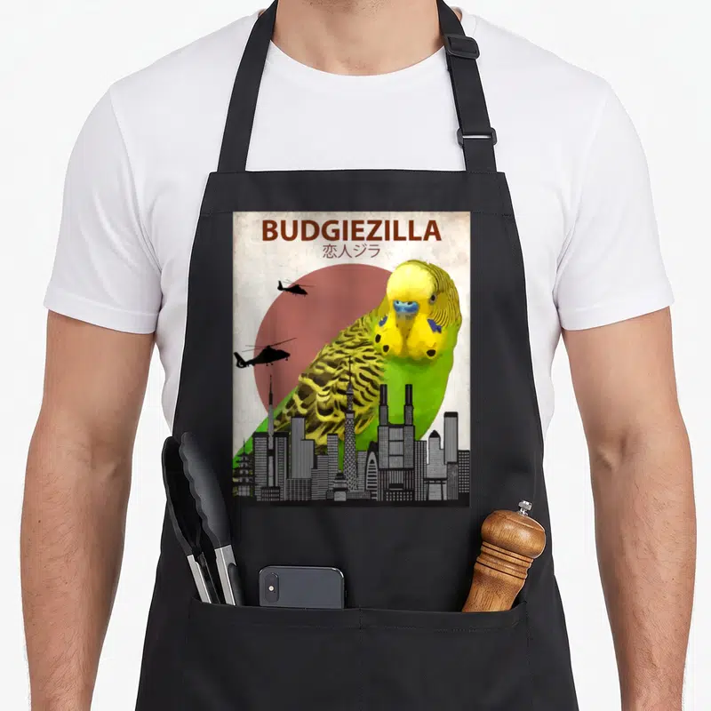 Budgiezilla parakeet lover apron - Giant yellow and green budgie looming over city skyline with helicopters flying — shown on a black cotton canvas apron against a white background