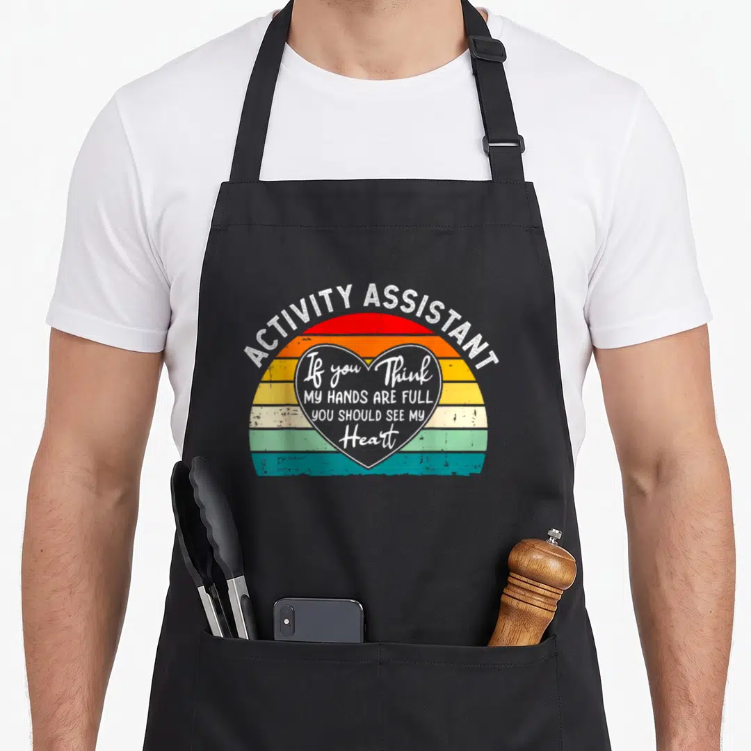 Hand activity apron - Retro striped sunset with white heart outline containing Activity Assistant text — shown on a black cotton canvas apron against a white background