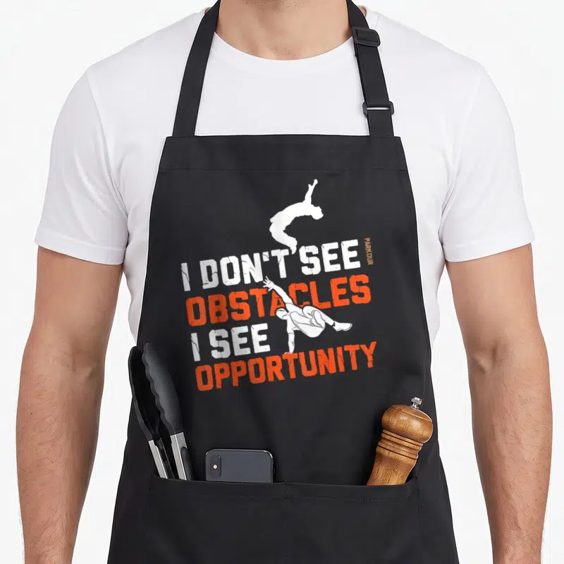 Free running parkour apron - Stacked I DON'T SEE OBSTACLES I SEE OPPORTUNITY text with two white leaping athlete — shown on a black cotton canvas apron against a white background