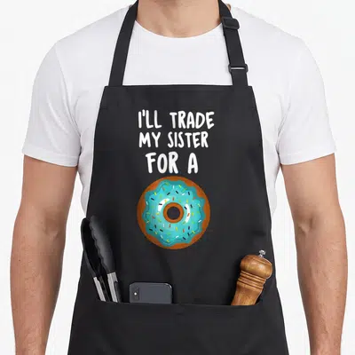 Ill donut apron - Cartoon blue frosted donut with sprinkles below white text reading I'll Trade My Sister — shown on a black cotton canvas apron against a white background