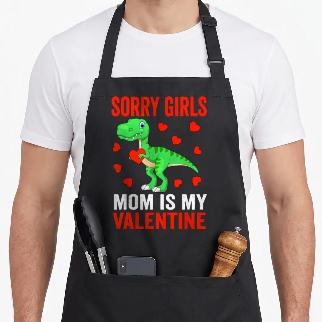 Sorry toddler valentine's day apron - Green cartoon dinosaur holding red heart with SORRY GIRLS MOM IS MY VALENTINE text — shown on a black cotton canvas apron against a white background