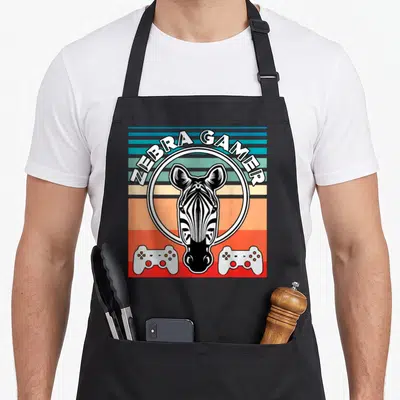Gamer zebra apron - ZEBRA GAMER text over zebra head and two controllers on colored stripes — shown on a black cotton canvas apron against a white background