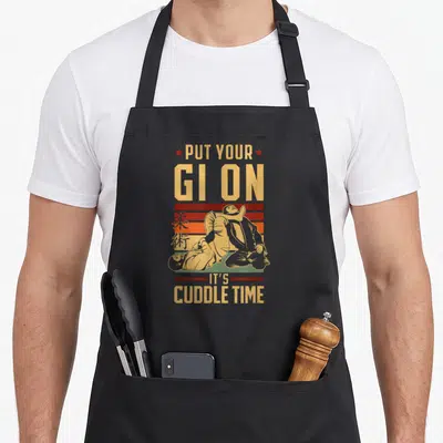Cuddle jitsu apron - Retro martial arts pun design with grappling figures and Japanese characters in yellow red orange — shown on a black cotton canvas apron against a white background