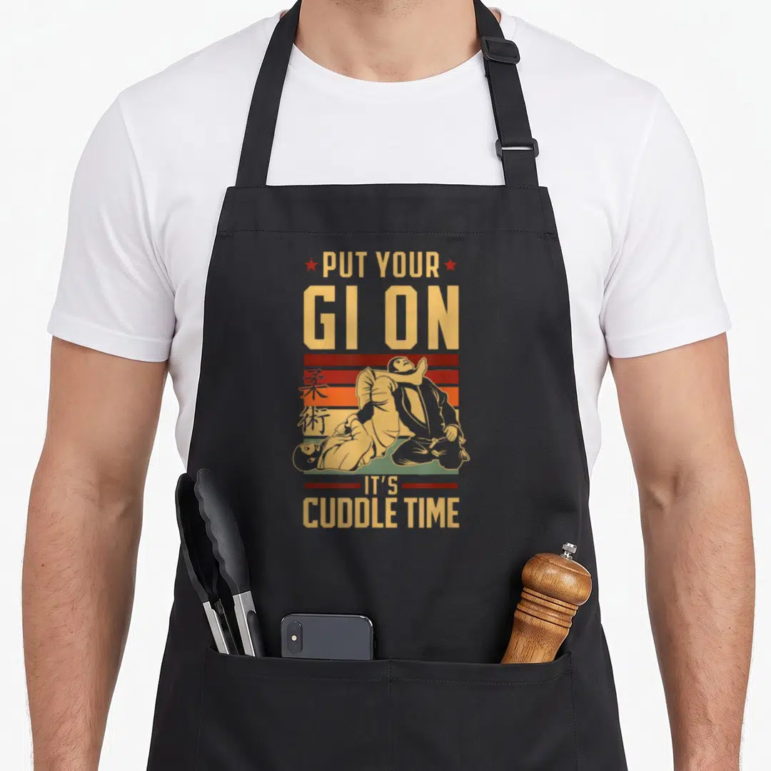 Cuddle jitsu apron - Retro martial arts pun design with grappling figures and Japanese characters in yellow red orange — shown on a black cotton canvas apron against a white background