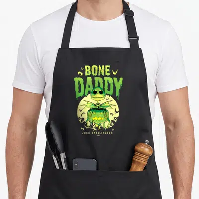 Bone daddy apron - Jack Skellington in pumpkin with dripping green bone daddy text and yellow moon — shown on a black cotton canvas apron against a white background