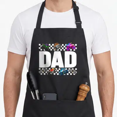 Family crew truck apron - DAD text with three monster trucks above and two below plus four checkered flags in cartoon — shown on a black cotton canvas apron against a white background
