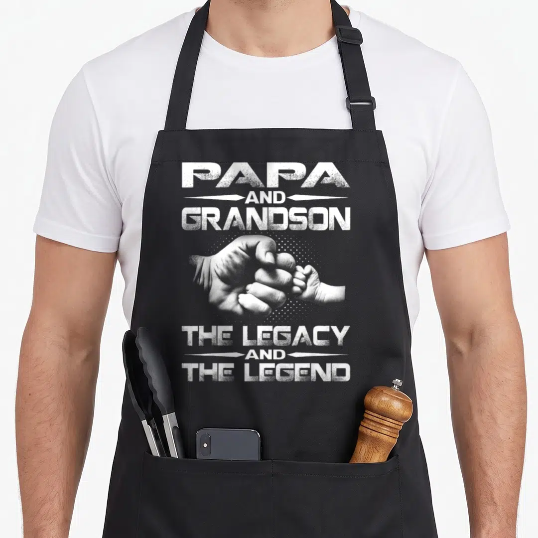 Grandson papa apron - White text reading Papa and Grandson The Legacy and The Legend over two fist bumping hands — shown on a black cotton canvas apron against a white background