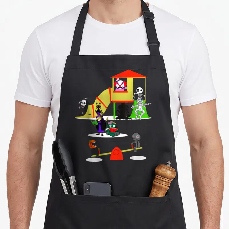 Icon horror movie apron - Cartoon monsters including a clown, skeleton, mummy, and witch playing on a colorful — shown on a black cotton canvas apron against a white background
