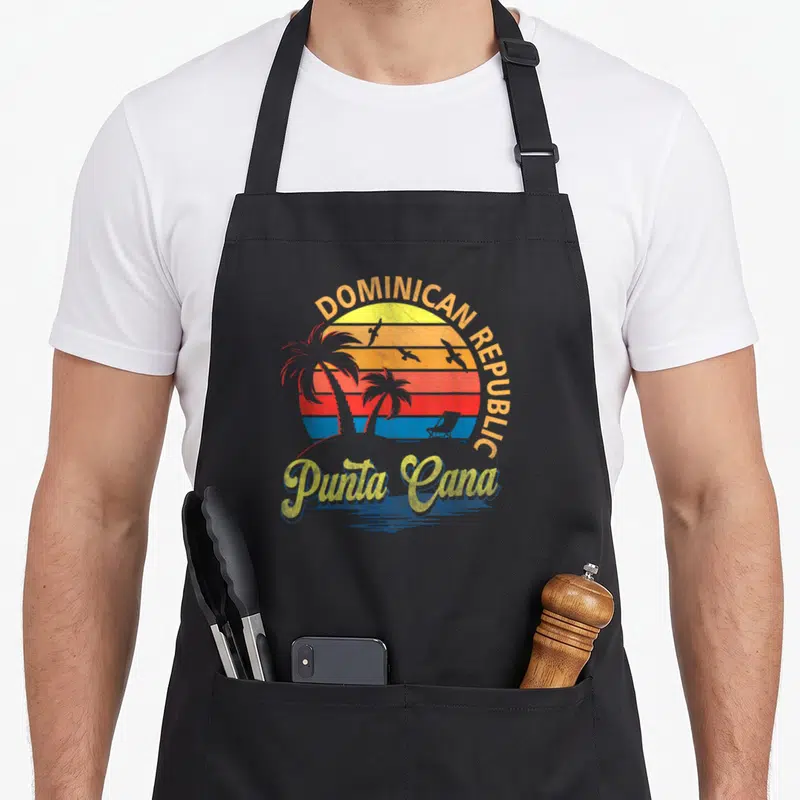 Sunset dominican apron - Retro sunset with DOMINICAN REPUBLIC text, Punta Cana script, and palm tree silhouettes — shown on a black cotton canvas apron against a white background