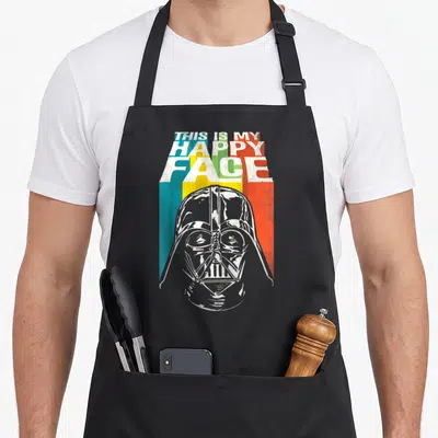 General face apron - Darth Vader helmet below stacked THIS IS MY HAPPY FACE text over three colored stripes — shown on a black cotton canvas apron against a white background