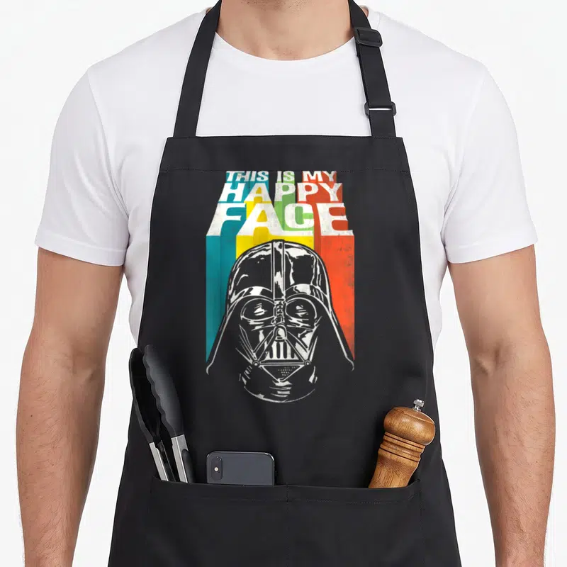 General face apron - Darth Vader helmet below stacked THIS IS MY HAPPY FACE text over three colored stripes — shown on a black cotton canvas apron against a white background