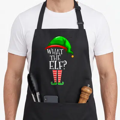 Elf christmas personalized family aprons - Cartoon elf hat and striped shoes framing stacked text WHAT THE ELF? — shown on a black cotton canvas apron against a white background