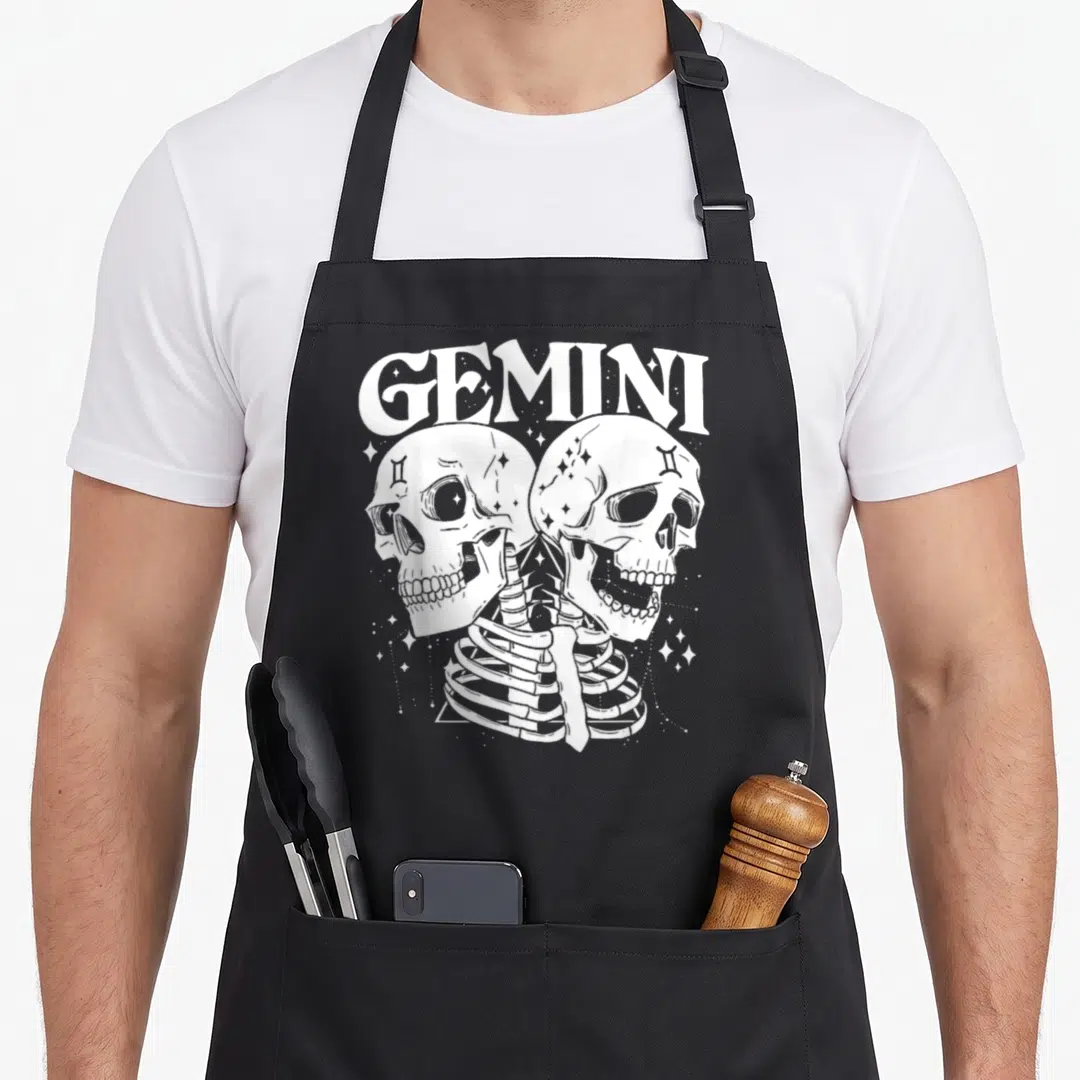 Gemini witch apron - White line art of two skulls facing a ribcage under arched GEMINI text with stars — shown on a black cotton canvas apron against a white background