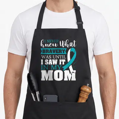 Bravery ribbon apron - Teal and white awareness ribbon overlaps stacked text reading I NEVER knew What BRAVERY WAS — shown on a black cotton canvas apron against a white background