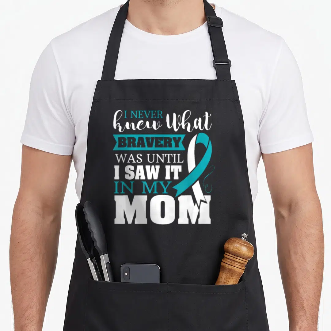 Bravery ribbon apron - Teal and white awareness ribbon overlaps stacked text reading I NEVER knew What BRAVERY WAS — shown on a black cotton canvas apron against a white background