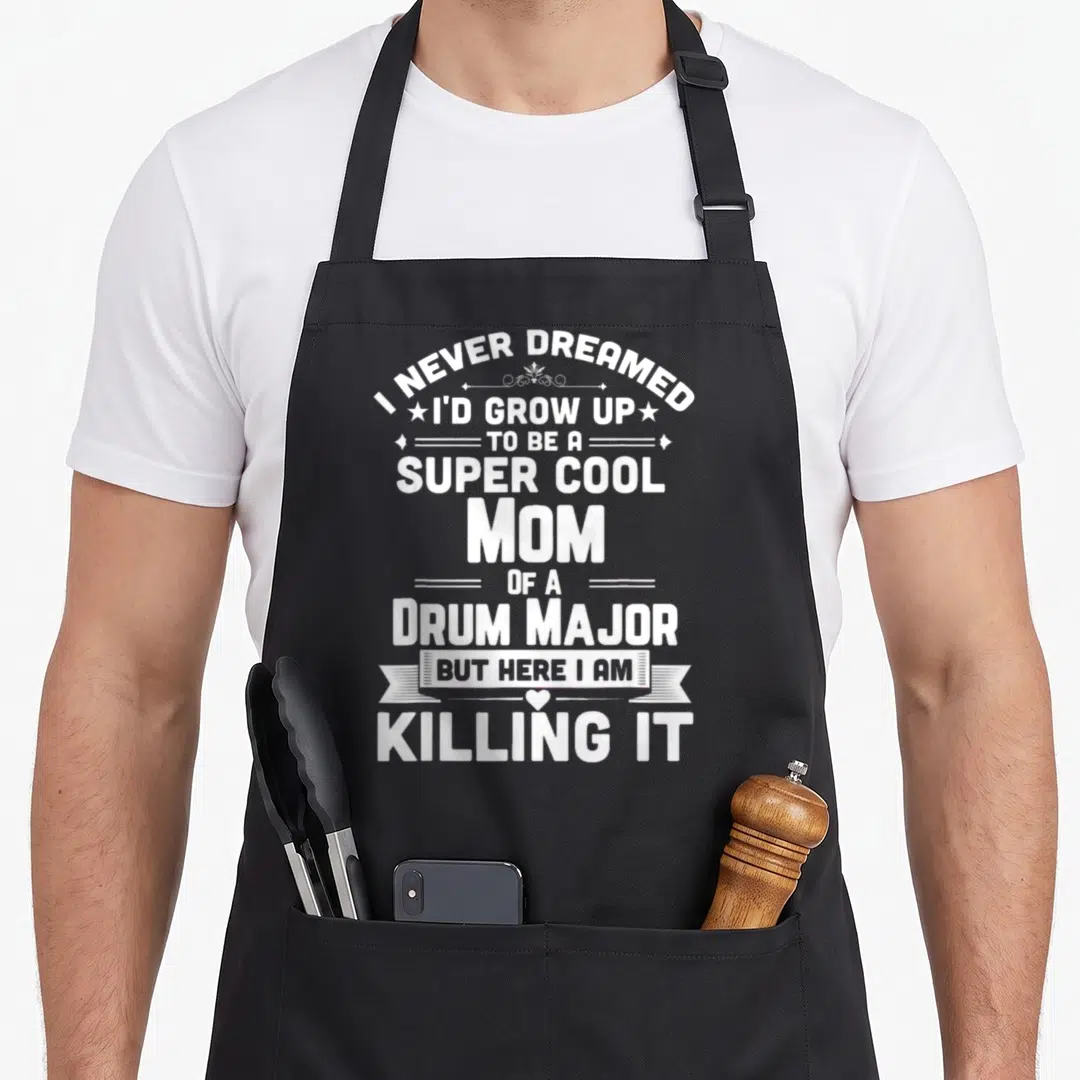 Band major apron - Stacked white typography reading I NEVER DREAMED TO BE A SUPER COOL MOM OF A DRUM MAJOR WITH BANNER — shown on a black cotton canvas apron against a white background