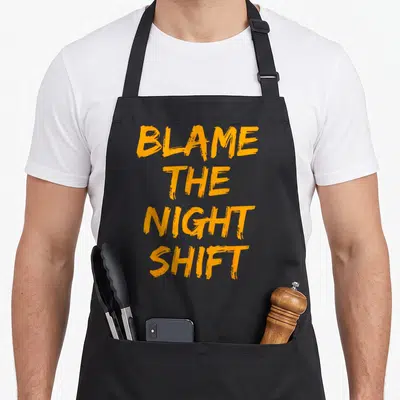 Shift swagazon apron - Orange stacked text reading BLAME THE NIGHT SHIFT in rough font — shown on a black cotton canvas apron against a white background